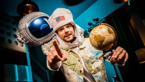 Een foto van Buddy die naar de camera wijst en in zijn andere hand een wereldbol houdt. Hij heeft een muts op die lijkt op de helm van een astronaut, met een vlag van de Verenigde Staten erop. Erachter is een ballon van een astronaut in pak te zien.