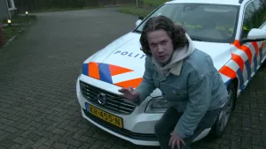 Een foto van Pascal die gehurkt staat voor een witte politieauto van het merk Volvo. Op de motorkap staat "Politie" en op de motorkap en zijkant staan blauwe en rode strepen. Er zitten twee agenten in, door de gele strepen op hun kostuum te zien.