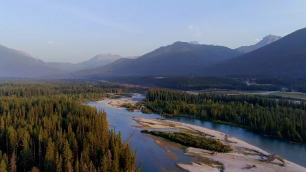 Een foto van een landschap met groene dennenbomen links en rechts van een rivier. In de rivier liggen stukken land met een ondergrond van zand. Achteraan zijn ongeveer zeven bergen of heuvels deels te zien.