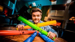 Een foto van Buddy achter een tafel met in zijn handen één oranje en één groene plastic buis of koker. Op de groene buis staat "Fa", op de oranje buis staat "E". Op de tafel liggen twee blauwe buizen, één met "Sol" erop, en één rode buis met "Do" erop.