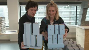 Een foto van een man en Janouk die achter een rechthoekig ding staan met hoge, rechthoekige, blokken erop. Het zijn blauwgrijze voorbeelden van een gebouw, maquettes. Bij één maquette staat een zwarte letter op ieder blok, een "W" een "K"  of een "H".