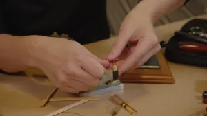 Een foto van de handen van iemand achter een lichtbruine tafel, waarop stukjes door midden gespleten bamboehout liggen en dubbelgevouwen bamboehout op een soort stokje. De persoon heeft een stukje opgevouwen bamboehout vast, dat op een soort stokje zit.