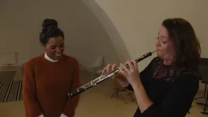 Een foto van een vrouw die speelt op een hobo, een zwart, houten blaasinstrument. Het is een lange buis met zilverkleurige knoppen en buisjes erop en er steekt een staafje uit, het mondstuk waar de vrouw op blaast. Sosha staat er met een blije lach naast.