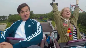 Een foto van Janouk en een man in een achtbaan die al een stukje omhoog is gegaan. Janouk zit rechts, kijkt blij met haar ogen dicht en heeft haar handen omhoog. De man links heeft zijn ogen wijdopen, zijn handen gevouwen en kijkt naar beneden.