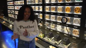 Een foto van Sosha die naast een muur staat in museum Micropia in Amsterdam waaraan ronde, verlichte schaaltjes met microben, zoals bacteriën en schimmels hangen. Er hangen naamplaatjes onder de microben en hoofdtelefoons om uitleg te krijgen.