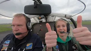 Een foto van Janouk en een man die in een rondom doorzichtig vliegtuigje zitten, dat nog op de landingsbaan staat of net begonnen is met opstijgen. Ze dragen een hoofdtelefoon met microfoon. Janouk houdt twee duimen omhoog en kijkt naar de camera.