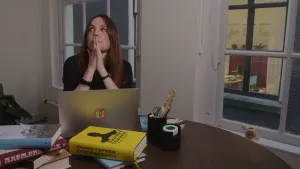 Een foto van dichter Ellen Deckwitz, die met haar handen tegen elkaar aan voor haar mond naar de linkerbovenhoek van de foto kijkt. Voor haar op tafel staat een laptop en liggen twee stapels boeken. Ellen denkt na tijdens het schrijven van een gedicht.