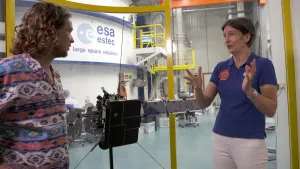 Een foto van Pascal en een vrouw met haar handen voor haar lichaam geheven. Ze staan naast een voorbeeld van een ruimtesonde, de Solar Orbiter. Het is een zwart kastje met drie lange antenes met gele bolletjes erop en aan twee kanten rechthoekige stukken.