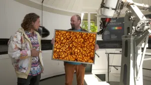 Een foto van Pascal die in een ronde ruimte met witte muren en plafond staat. Rechts staat een grote telescoop om naar de sterren te kunnen kijken. Naast Pascal staat een man met een foto van het  oranjerode oppervlak van de zon met bruine lijnen erdoor.