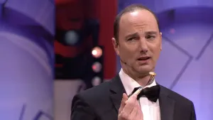 Een foto van een presentator in een zwart pak, met een witte blouse eronder en een strikje om zijn nek. De presentator heeft een vork vast met een ovaal stukje lichtbruin eten erop geprikt. Hij kijkt naar links op de foto.