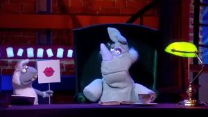 Een foto van de pop van Nico de Neushoorn met een bril. Hij zit op een groene stoel achter een bureau met een boek erop. Naast pop Nico staat de pop Stella Schaap. Stella heeft een bord vast met getekende roze lippen erop.