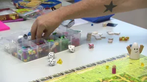 Een foto van een plastic bakje met vakjes vol kleurige pionnen, vierkantjes en dobbelstenen waar een hand iets uitpakt. Ervoor liggen vijf dobbelstenen en een bordspel met groene vlakken met een pion, twee gele stukjes en een soort lachende eivorm erop.