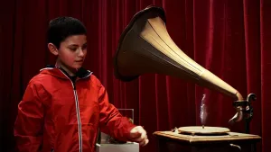 Een foto van Fernando naast een platenspeler met een grote hoorn en een ronde draaischijf, waar een plaat met muziek opgelegd kan worden. Er ligt geen plaat op, maar erboven is een driedimensionaal voorbeeld van het muziekinstrument viool te zien.