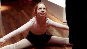 Een foto van Sarah in een soort ballet of danskleding, die met naar de zijkant gestrekte benen en armen op de grond zit. Ze kijkt omhoog naar iemand waarvan alleen een zwarte vorm van een jas of kledingstuk zichtbaar is aan de rechterkant. 