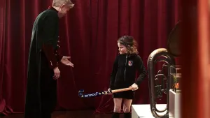 Een foto van Charlotte in hockeykleding met een hockeystick. Een man in een donkergroene Jas met goudkleurige knopen en rode kleding eronder wijst met zijn hand naar de hockeystick. Een blaasinstrument met een ronde opening staat op een witte tafel.