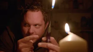 Een foto van Diederik met een aansteker in zijn hand. Hij drukt op het knopje van de aansteker, en er komt een vlam uit de bovenkant. Rechtsvoor staat een brandende witte kaars. Diederik kijkt naar links naar iets buiten beeld.