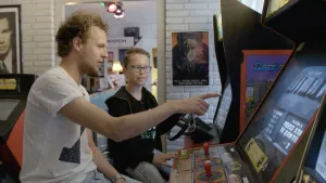 Een foto van Tim en Tristan, die achter een arcadekast zitten. Dat is een kast om een computerspel te spelen met twee stuurknuppels, knoppen en een beeldscherm, waar Tim naar wijst. Op het scherm staat "Ultimate", het spel is Ultimate Mortal Kombat 3.