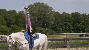 Een foto van Rosaline die bezig is een koprol te maken op een wit paard, dat buiten loopt over een zandvlakte. Rosaline haar benen staan omhoog in de lucht en ze heeft twee zwarte handvaten vast die op de rug van het paard vastgezet zijn. 