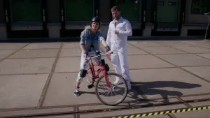 Een foto van Janouk die buiten op een rode fiets zit en een zwarte helm, polsbeschermers en kniebeschermers draagt. Ernaast staat prof in een wit werkpak. Ze kijken naar de camera.  Ze staan achter een bedrijfsgebouw op een verder lege weg.