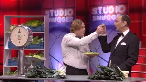 Een foto van een tafel met stronken van de groente prei erop en een weegschaal. Erachter staat een kast met blauwe mandjes vol groenten, sla en bloemkool. Achter de tafel staat een man met een bord spruitjes, die een spruitje geeft aan een presentator. 