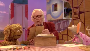 Een foto van meneer Aart die naar een bruine kaart in zijn handen kijkt die hij uit een houten bak op tafel heeft gehaald. Tommy en Ieniemienie zitten ieder aan één kant ernaast, met op de tafel voor hen een tekening op tekenpapier, stiften en potloden.