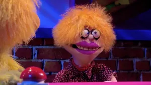 Een foto van pop Annewil Stoeien. De pop heeft oranjeachtig haar, dikke lippen met paarse lippenstift, schele, ronde ogen en een openstaande mond. De pop draagt een donkere blouse en een rode ketting. Links is een rode drukknop te zien naast Opper de Pop.