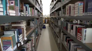 Een foto met aan twee kanten metalen rekken, waarvan de planken vollliggen met boeken. Op de boeken zitten stickers met logo's en een Letter op de zijkant. Op de linkerplank is de letter "F" voor de plank te zien. De rekken staan in een bibliotheek.