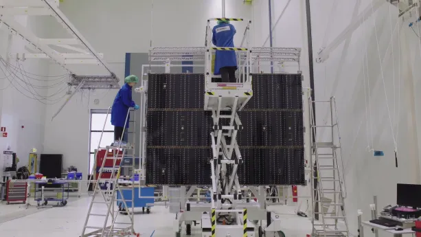 Een foto van een witte ruimte met twee mensen die een blauwe jas, handschoenen en een haarnetje dragen. De mensen staan op een ladder en een soort lift. Voor hen staat een zwart, rechthoekig gedeelte van een satelliet, die ze aan het testen zijn. 