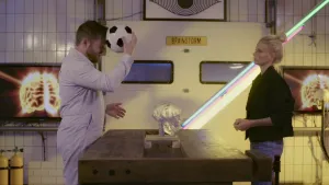 Een foto van prof en Janouk achter een werkbank, dat is een soort tafel met een klem eraan, waarop je iets vast kunt draaien. In het midden van de tafel staat een doorzichtig voorbeeld van een menselijke schedel. Prof houdt een voetbal tegen zijn hoofd.