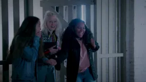 Een foto van Janouk die lachend naar de camera kijkt en twee kinderen voor Janouk die ook lachen. Ze staan in een gebouw met witte stenen en voor een geopende, witte deur van houten planken met een wit, houten hek van planken eromheen.