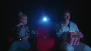 Een foto van Janouk en prof die op rode stoelen zitten in een donkere bioscoopzaal. De stoel ertussen is leeg. Erachter is het lichtblauwe licht van de projector te zien. Prof heeft een bak op zijn schoot met "Popcorn" erop en hij eet een stukje popcorn.