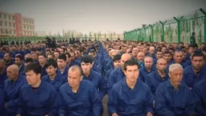 Een foto van een groep mannen op een plek in de buitenlucht, afgezet met groene hekken en bewaakt door soldaten die achteraan staan. De mannen zitten op de grond en dragen allemaal een blauw pak. Het zijn Oeigoeren in een 'heropvoedingskamp' in China.