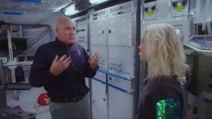 Een foto van astronaut André Kuipers en Janouk. Ze staan in een ruimte met rechts witte platen met schroefjes erop. Aan één deel zitten twee blauwe, metalen buizen om je aan vast te houden. Achteraan hangen kleine kastjes , sommige met draadjes eraan.