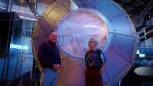 Een foto van Janouk en astronaut André Kuipers die naar de camera kijken. Ze staan voor een groot, rond ding met een doorzichtige koepel in het midden. Aan de zijkant van het ding zitten gele buizen, ene soort handvaten. 