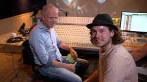 Een foto van Pascal en een man achter een groot mengpaneel met knopjes en schuifjes in een geluidsstudio. Aan de rechterkant staat een computerscherm. De man heeft twee lichtgroene trechters vast in zijn handen. Pascal zit ernaast en kijkt naar de camera.