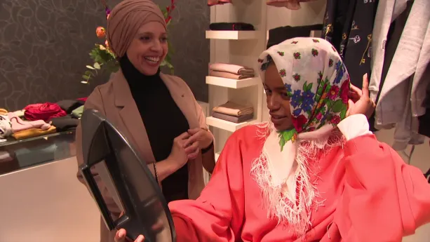 Een foto van een mevrouw met een lichtbruine hoofddoek om. Eva heeft ook een hoofddoek om, een witte met blauwe en paarse bloemetjes erop. Eva draagt een wijd rood pak en heeft een ronde spiegel vast. Ze kijkt in de spiegel hoe de hoofddoek zit.