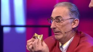 Een foto van een man die zijn vingers gestoken heeft in een gevouwen ding van origami papier, waarvan twee puntjes omhoogsteken. Het papier heeft een goudkleurige glans. Het gevouwen ding is een happertje, waarmee je een vingerspel kunt spelen.