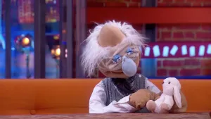 Een foto van pop professor Bolleboos met grijs haar, een rimpelig gezicht, een lichtblauwe neus en een bril. De pop heeft een roze knuffelkonijn vast en kijkt ernaar. Hij zit op een bruine bank achter een bruine tafel.