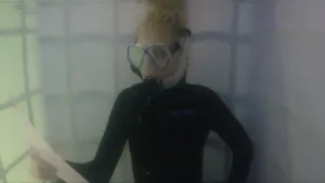 Een foto van Janouk in een soort bak met water. Ze heeft een donkergekleurd pak aan en draagt een blauwe en deels doorzichtige duikbril, waarvan een doorzichtige, plastic slang loopt naar een zwart mondstuk. Ze leest van een wit vel papier in haar hand.