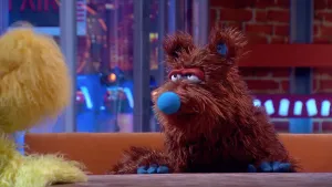 Een foto van pop Berend B. met een bruine, langharige vacht, een blauwe neus en blauwe vingers of klauwen. De wenkbrauwen van de pop hangen deels voor de ronde ogen. De pop zit op een bruine bank achter een bruine tafel. Links in beeld zit Opper de Pop.