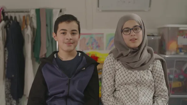 Een foto van Joussef en Imane. Joussef draagt een donkerblauw jasje met zwarte mouwen, Imane een grijze blouse met ronde vormen en een lichtbruine hoofddoek. Erachter staan bakken met speelgoed, hangen tekeningen aan de muur en kleding aan een rek.