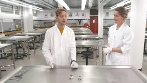 Een foto van Elbert en een mevrouw in een witte jas en witte plastic handschoenen achter een stalen snijtafel. Op de tafel liggen mesjes en scharen en in het midden twee koeienogen. Elbert kijkt met grote ogen naar beneden naar een plek achter de camera.