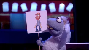 Een foto van pop Stella Schaap met een bord waar een getekende man op staat met rood haar, een rode baard, een grijze blouse en een blauwe broek. Stella kijkt naar het bord en heeft een witte vacht, grijs bij kop en poten, en vooruitstekende, gele tanden.