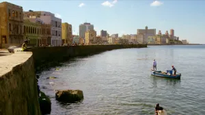 Een foto van een stad in Cuba met allemaal gebouwen van verschillende kleuren links en rechts een hoge kade met een kleine houten boot met twee staande mensen erin. Aan de kade zitten mensen met hun benen over de rand van de kade naar het water te kijken.
