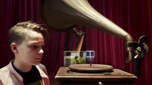 Een foto van Ilya die naast een ouderwetse platenspeler met een grote hoorn erop staat.  Op de platenspeler ligt geen plaat, maar uit het ronde gedeelte waar de plaat normaal ligt, komt een soort projectie van een kubus met lichtgevende lijnen.