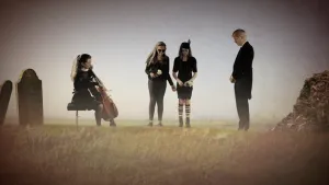 Een foto van vier mensen in zwarte kleding op een begraafplaats naast twee grafstenen met tekst. Een vrouw speelt met een strijkstok op een snaarinstrument, een cello. Twee meisjes hebben een witte bloem vast en een man staat ernaast met gevouwen handen.