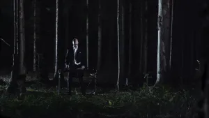 Een foto van een man in een zwart pak die in een donker bos tussen de bomen staat. Voor de man staat een instrument, een xylofoon. Dat is een soort piano met toetsen waar je op moet slaan. De man speelt op de xylofoon met houten stokken.