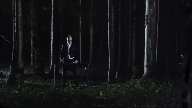 Een foto van een man in een zwart pak die in een donker bos tussen de bomen staat. Voor de man staat een instrument, een xylofoon. Dat is een soort piano met toetsen waar je op moet slaan. De man speelt op de xylofoon met houten stokken.