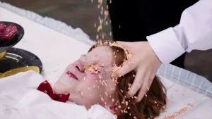 Een foto van een jongen die op wit kleed op een tafel ligt. Onder zijn gezicht is een stukje van zijn witte blouse en rode strik te zien. Een andere jongen houdt met één hand zijn hoofd vast en vruchtenhagel valt van boven op het gezicht van de jongen.