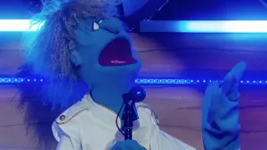 Een foto van pop Toontje Hoger die in een microfoon zingt. De pop heeft een blauwe huid en lichtbruin haar met kleine krullen. Toontje kijkt omhoog en houdt zijn hand voor zich omhoog. Hij draagt een witte jas met knopen op de schouders.